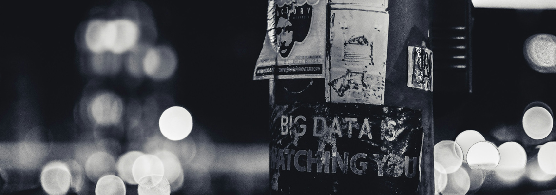 Das Bild zeigt eine Laternenpfahl bei Nacht, der mit verschiedenen Plakaten und Aufklebern beklebt ist. Im Fokus steht der Text "BIG DATA IS WATCHING YOU," der auf einem Aufkleber zu sehen ist. Das Bild ist in Schwarz-Weiß gehalten, was eine düstere und nachdenkliche Atmosphäre erzeugt.